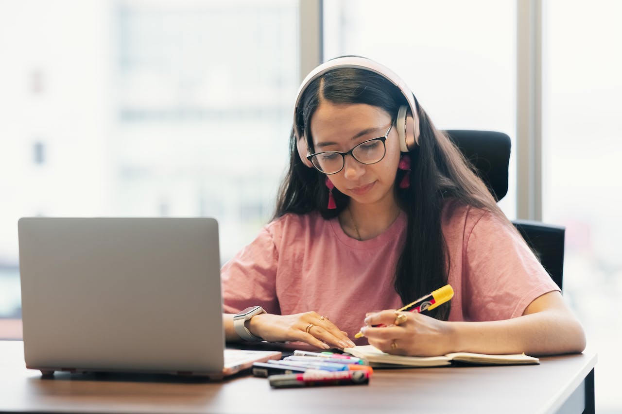 Young woman wearing headphones studying at desk with laptop and notebook.